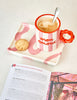 Que Rico Miguel Mon Amour mug filled with coffee on a pink patterned tray with a biscuit